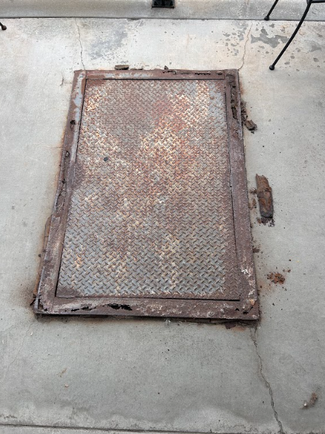 Rusted diamond plate access hatch and frame set in concrete, assessed for repair.